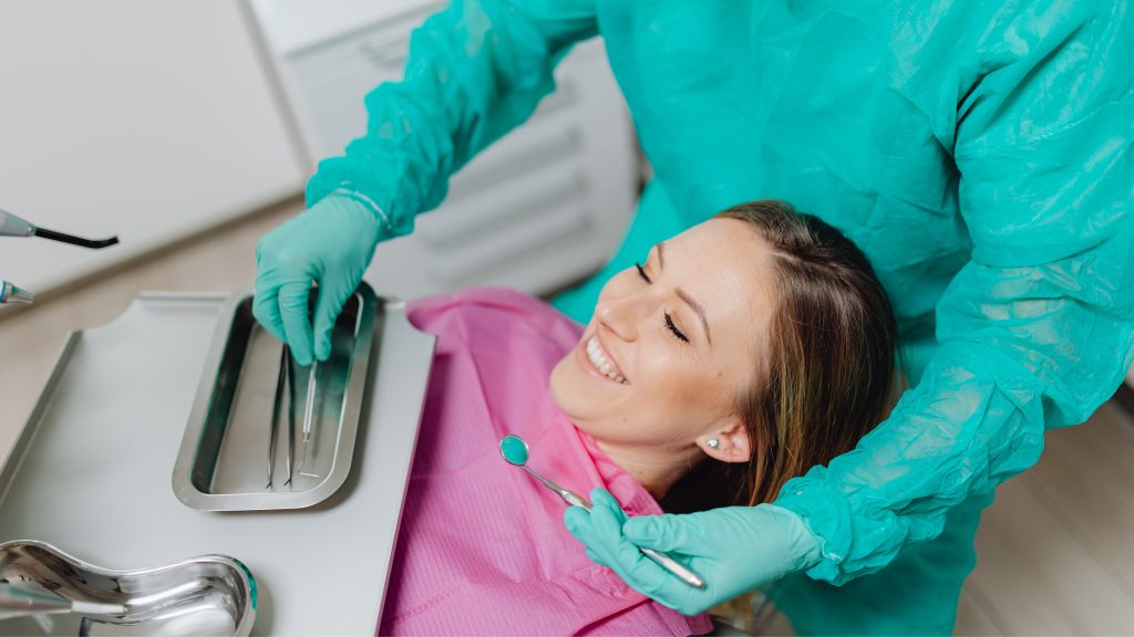 A smiling female patient lying in a dental chair while a dentist in a teal scrub gown prepares instruments.